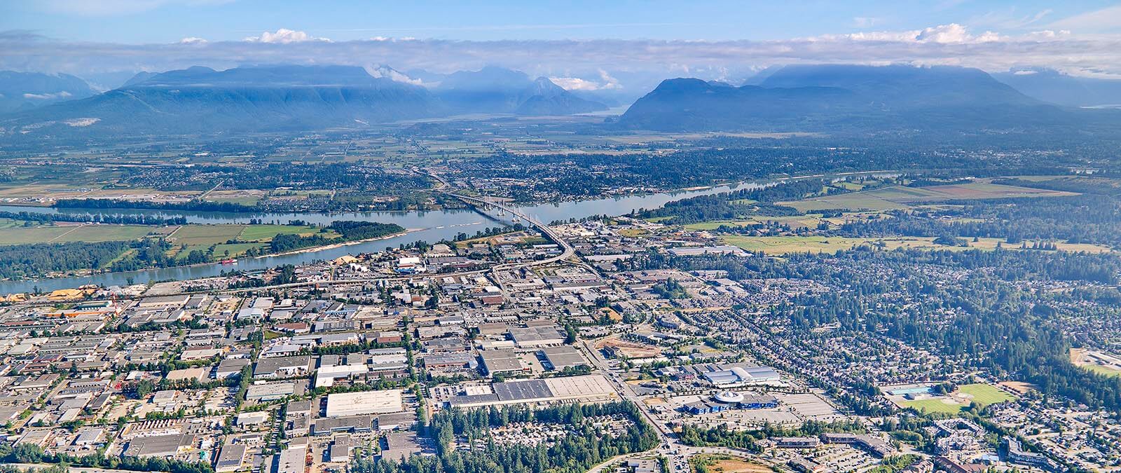 Aerial photo taken on a sunny day, trees and grass are green. Port Kells industrial area is in the foreground, the Fraser River and Golden Ears Bridge in the mid-ground, Golden Ears Mountains (and other mountains) in the background.